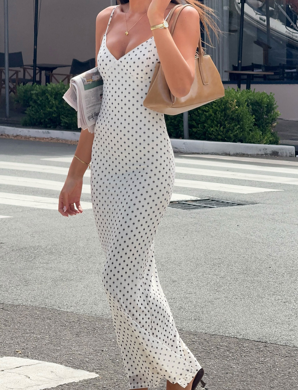 A woman in the TIGERMIST JULIANNA MAXI DRESS - POLKA DOT, featuring adjustable spaghetti straps, walks on a city street with a tan handbag and a newspaper under her arm.