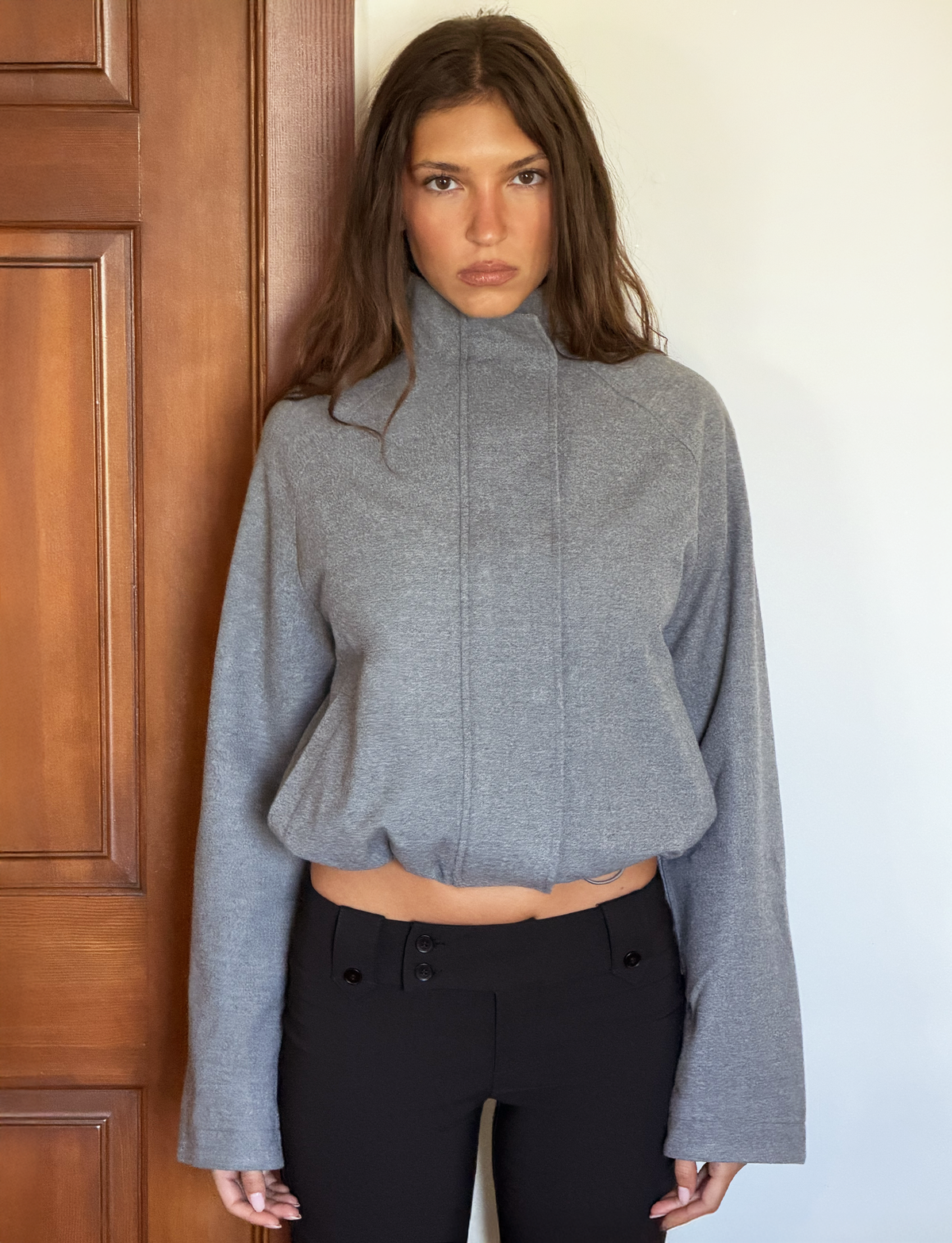 A woman with long brown hair stands indoors by a wooden door and white wall, wearing the TIGERMIST LENNEX JACKET in heather charcoal and black pants. She looks serious, her arms at her sides.