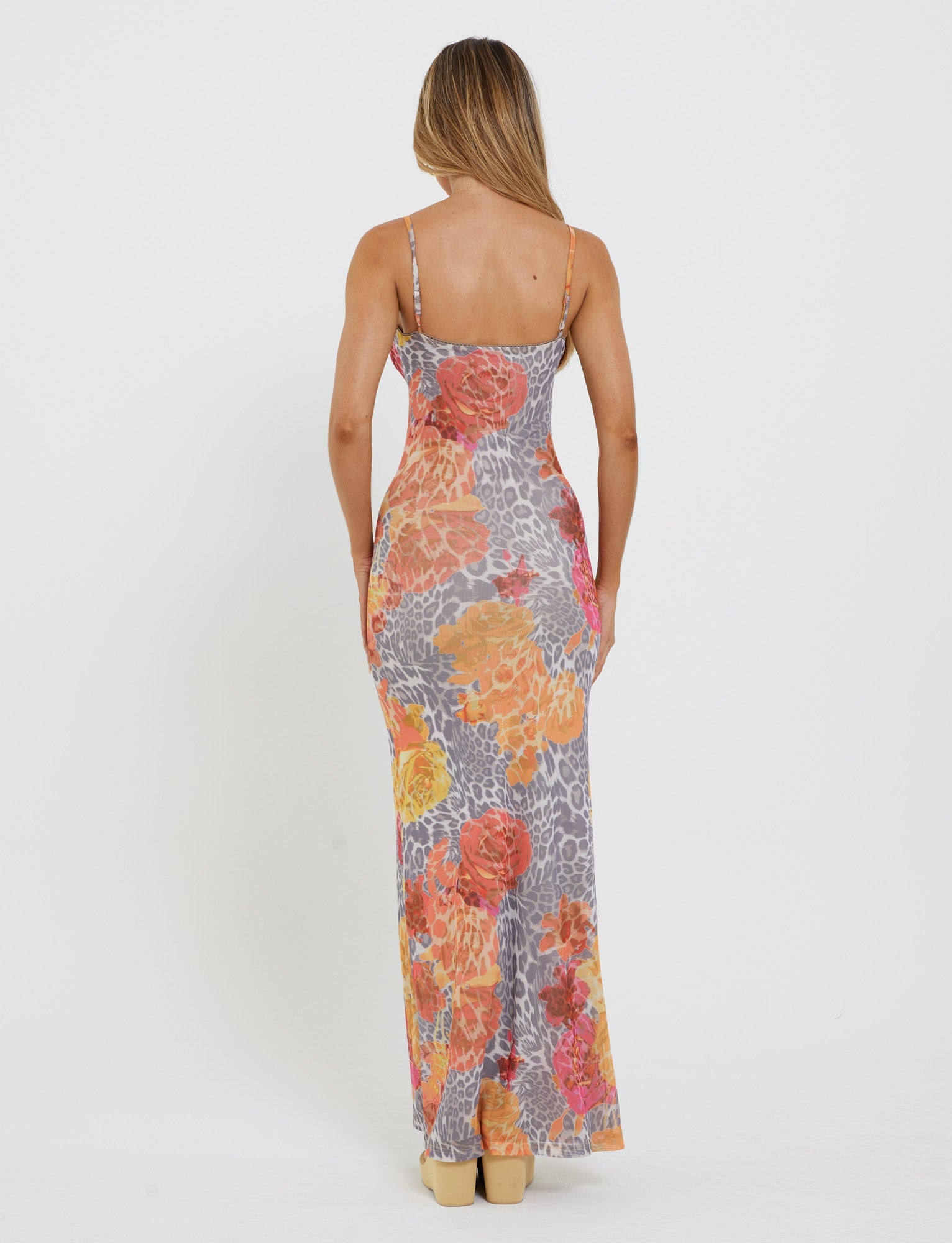 A woman with long hair, facing away from the camera, is wearing the Julianna Dress by TigerMist. The colorful sleeveless maxi dress features an abstract mix of floral and orange leopard print designs. She stands in a relaxed pose against a plain white background.
