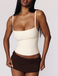 A woman with long dark hair wears the TIGERMIST MIDORI BANDAGE TOP in cream, featuring a fitted silhouette, sleeveless design, and square neckline, paired with a short brown skirt against a plain light background.