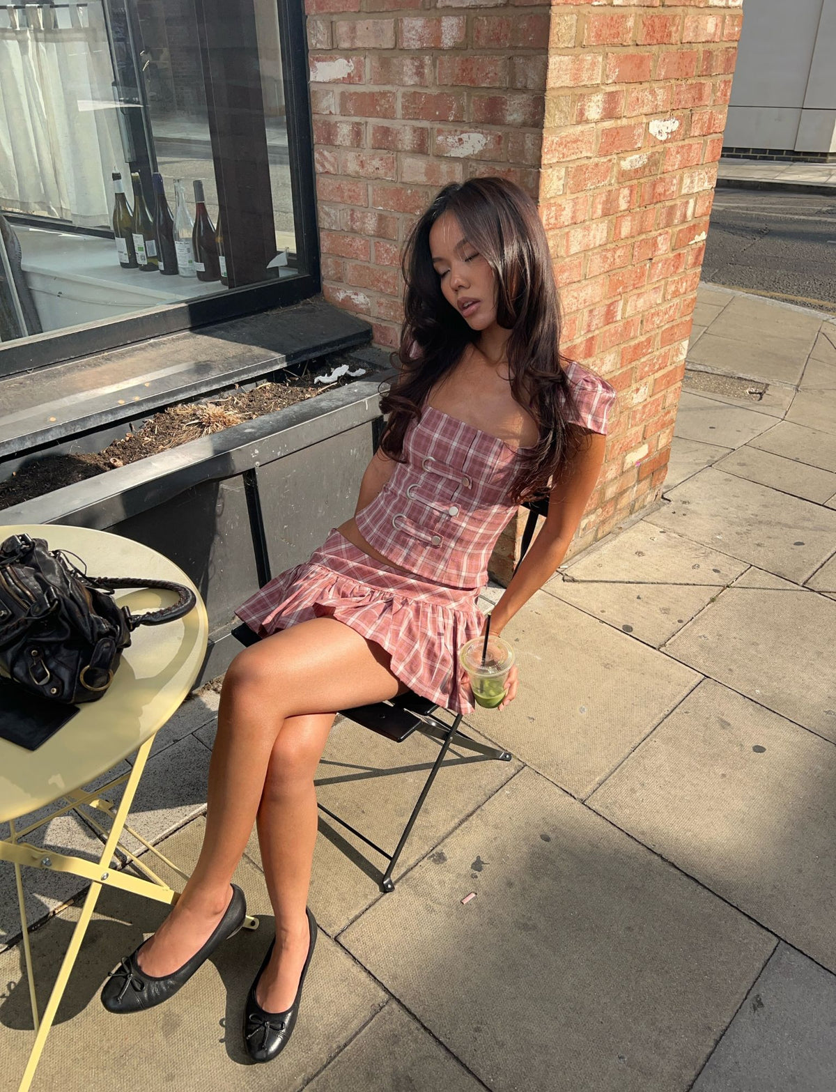 Wearing the TIGERMIST MIKA SKORT in pink plaid, a woman sits outside on a chair at a small table, enjoying a drink and soaking up the sunlight. A black purse is placed on the table beside her.