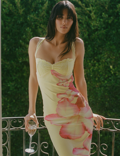 A woman in the TIGERMIST NYOMI MAXI DRESS - YELLOW HIBISCUS stands at a balcony railing with a glass of white wine, lush green foliage behind her, as she looks directly at the camera.