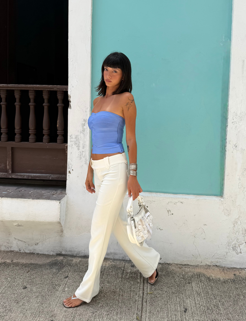 A woman with dark hair walks on a sidewalk in front of a turquoise wall and wooden railing, carrying a white purse. She wears the TIGERMIST NIYA BANDAGE TOP in cornflower blue, featuring a strapless, sweetheart neckline that pairs stylishly with her white pants.