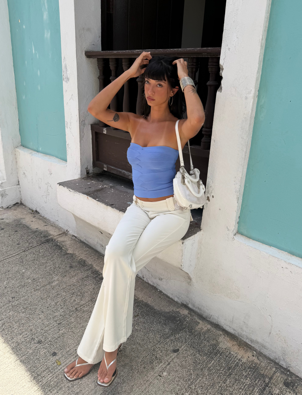 A woman sits on a windowsill outdoors, wearing the TIGERMIST NIYA BANDAGE TOP in cornflower blue—an elegant strapless top with a sweetheart neckline—paired with white pants. She holds her hair and accessorizes with a white bag, white sandals, and silver jewelry. The wall behind her features turquoise panels against a white backdrop.