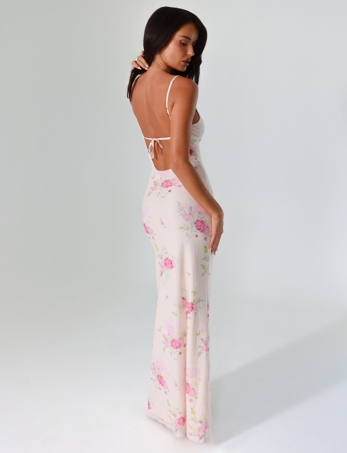 A woman models the OSIRIS DRESS from TIGERMIST, featuring a watercolor floral print in pink. The maxi dress has a low back and spaghetti straps. She poses against a plain white backdrop, gazing over her shoulder with her arms resting by her sides.
