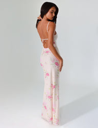 A woman models the OSIRIS DRESS from TIGERMIST, featuring a watercolor floral print in pink. The maxi dress has a low back and spaghetti straps. She poses against a plain white backdrop, gazing over her shoulder with her arms resting by her sides.