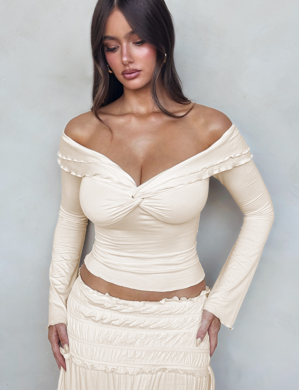 A woman wears the TIGERMIST DENALI LONG SLEEVE TOP - WHITE : BONE, featuring a sweetheart neckline and twist front, paired with a matching skirt. She stands against a light gray background, looking down with her hands at her sides.