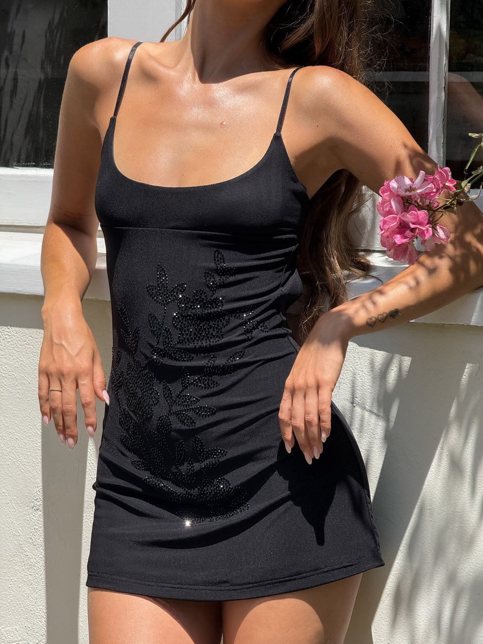 A woman stands in sunlight by a white wall, holding pink flowers, wearing the TIGERMIST OCTAVIA MINI DRESS - BLACK with adjustable spaghetti straps and floral embellishments. Her face is not visible.