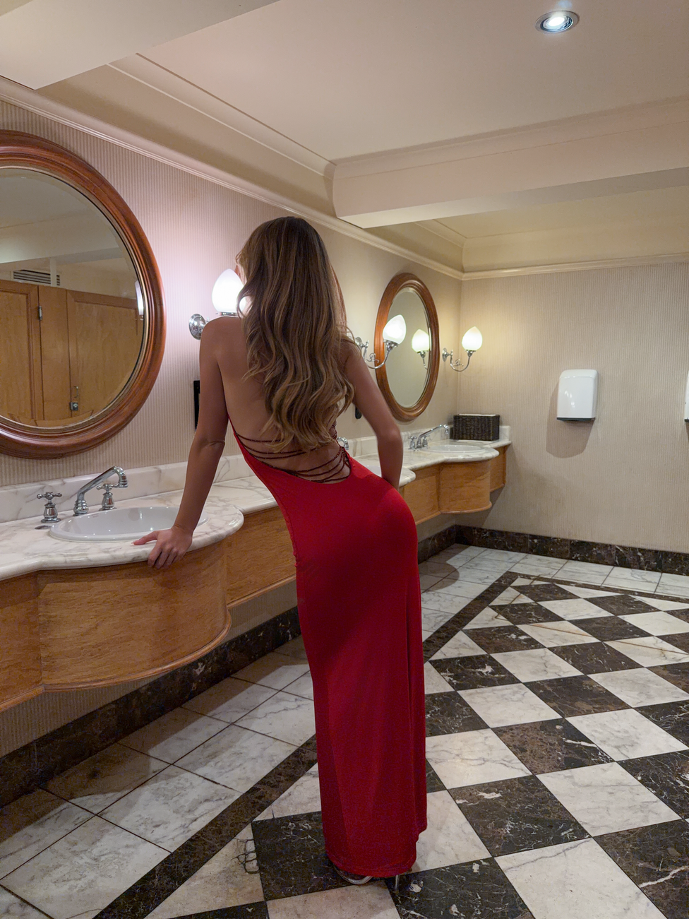 A woman wearing the TIGERMIST SOLYNE MAXI DRESS in red, featuring an open back, stands at a marble sink in a stylish bathroom with checkered floors and wood-trimmed mirrors, softly lit. She faces away from the camera.