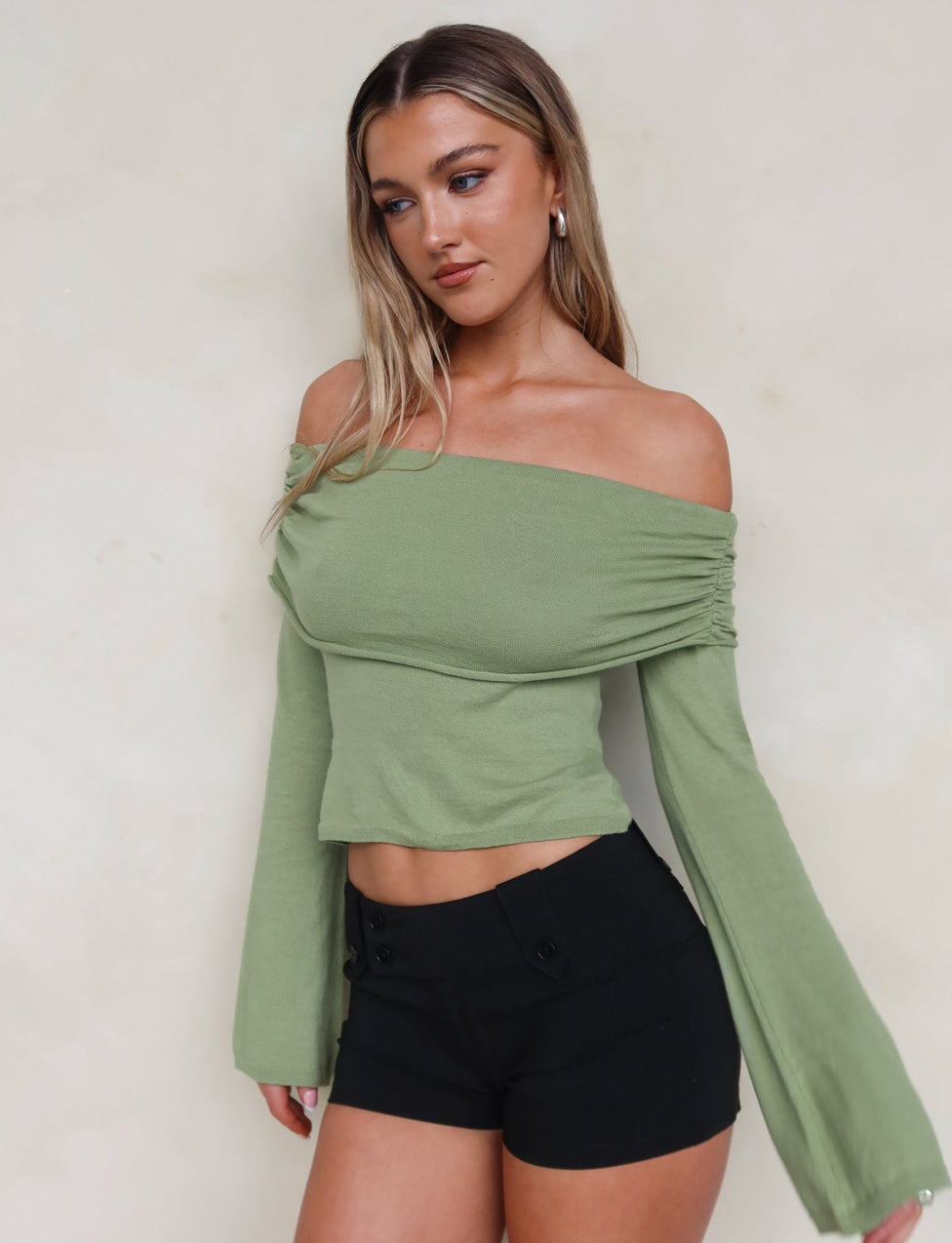 A woman with long, wavy blonde hair is looking to the side. She is dressed in the DIEDRE JUMPER - GREEN by TIGERMIST, an off-the-shoulder, long-sleeved, light green high stretch cotton top paired with high-waisted black shorts. The background is plain and light-colored.