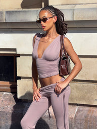 A woman stands outdoors against a stone wall, sunlight casting shadows as she wears sunglasses and the mauve CL - HALONA TOP from Tiger Mist USA with matching pants. She carries a brown handbag on her shoulder.