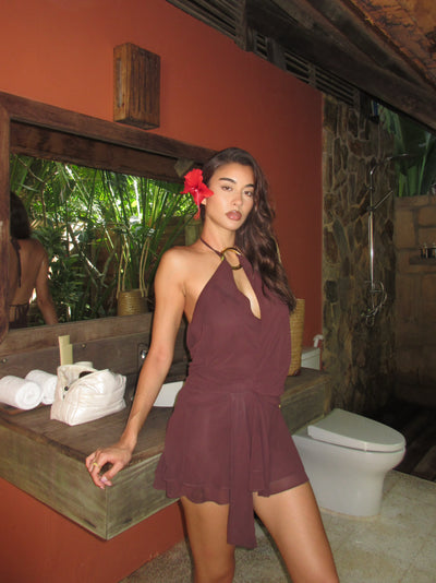 A woman wearing the TIGERMIST INDIGO MINI DRESS 2.0 in Chocolate poses confidently near a sink and mirror in a rustic bathroom with stone and wood accents, while lush greenery can be seen outside the window.