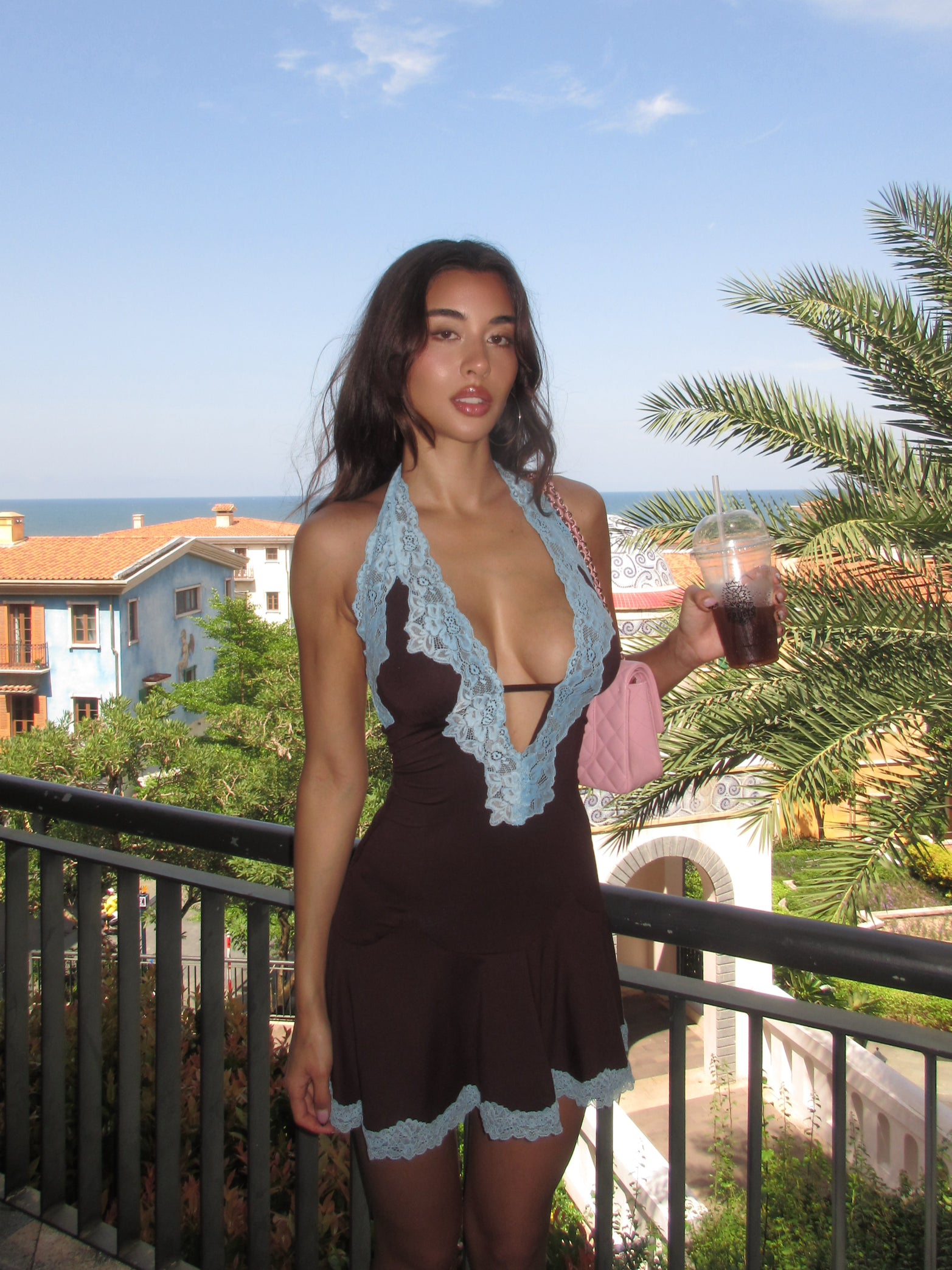 A woman in the TIGERMIST TALIYAH MINI DRESS - CHOCOLATE stands on a sunny balcony with an iced drink, overlooking Mediterranean buildings, palm trees, and the ocean.