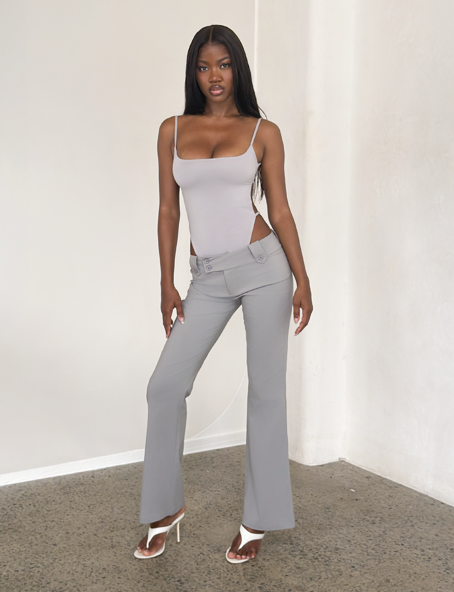 A woman poses confidently indoors against a white wall, wearing a fitted light gray tank top and the TIGERMIST KITTIE PANT - FROST GREY, featuring hip cutouts. She completes the look with white heeled sandals and loose dark hair.