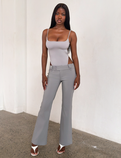 A woman with long, straight hair and a neutral expression stands indoors against a white wall, wearing the TIGERMIST KITTIE PANT - FROST GREY in stretch suiting fabric with side cutouts, paired with a matching tank top and white heeled sandals.