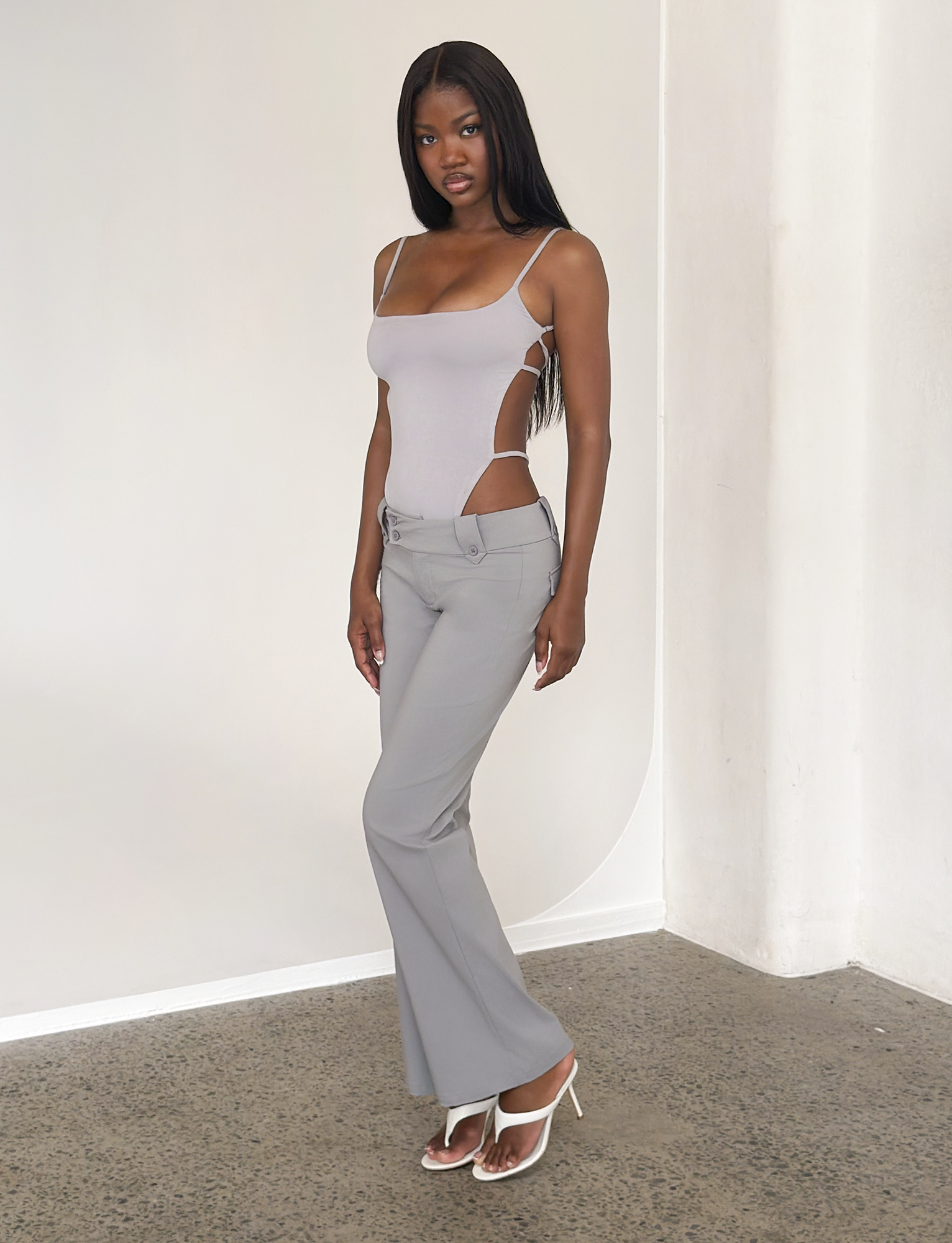 A woman with long straight hair stands indoors against a white wall, wearing a light gray fitted bodysuit with cutouts and the TIGERMIST KITTIE PANT in frost grey stretch suiting, paired with white heels and a neutral expression.