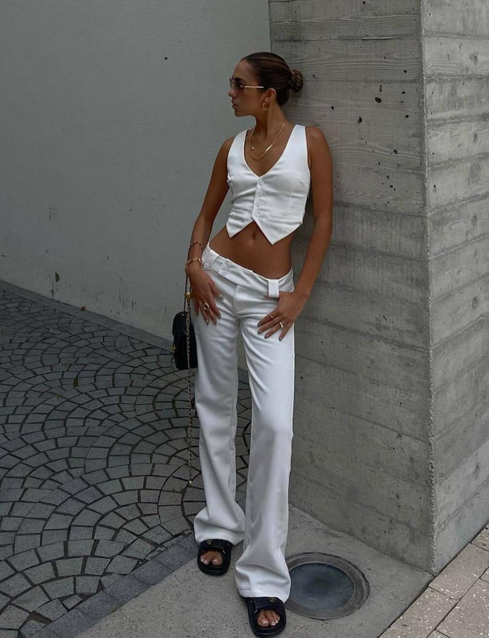 A woman poses outdoors in TIGERMIST's KITTIE TWILL PANT - WHITE, styled with a matching sleeveless vest, black sandals, sunglasses, and gold jewelry as she stands against a concrete pillar on a tiled sidewalk.