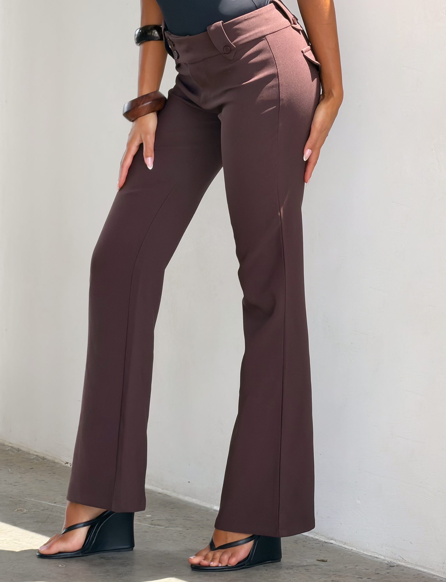 A person wears TIGERMIST's KITTIE PANT - BROWN : CHOCOLATE with a high waist and asymmetrical waistband, paired with black open-toed wedge sandals, standing against a white wall with one hand resting on their thigh.