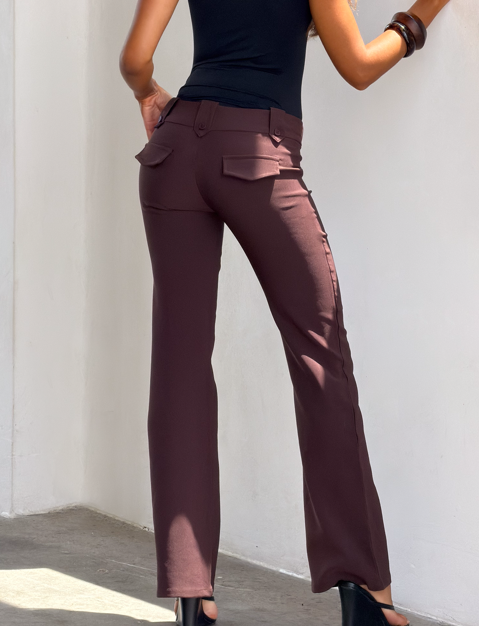 A woman in a black sleeveless top and TIGERMIST KITTIE PANT - BROWN : CHOCOLATE with an asymmetrical waistband leans against a white wall, one hand on her hip and the other on the wall. She wears black heels and a large brown bracelet. Photo taken from behind.