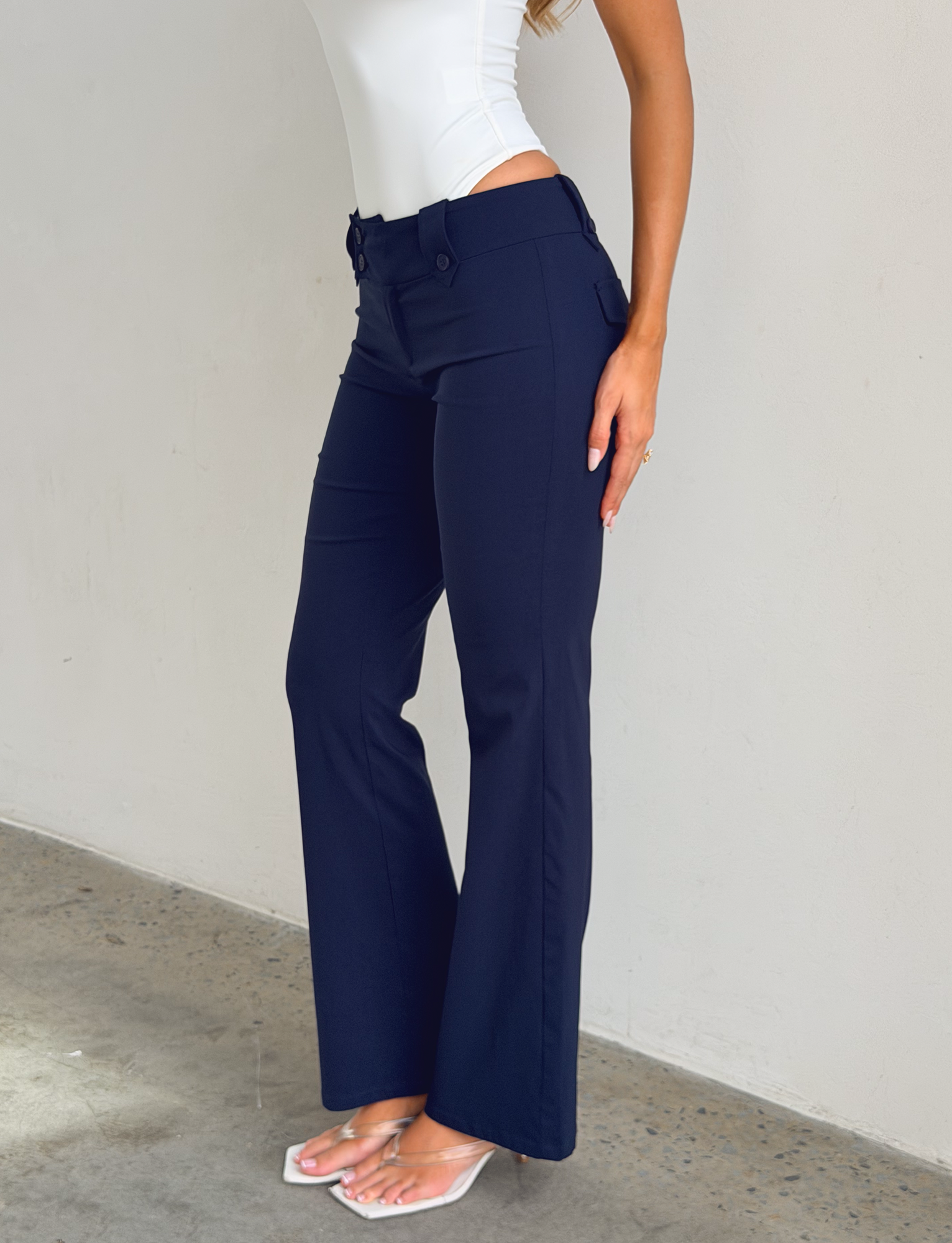 A person stands indoors by a light wall, wearing TIGERMIST's KITTIE PANT - NAVY in stretch suiting fabric, a white sleeveless top, and clear heeled sandals. Their face is not shown.