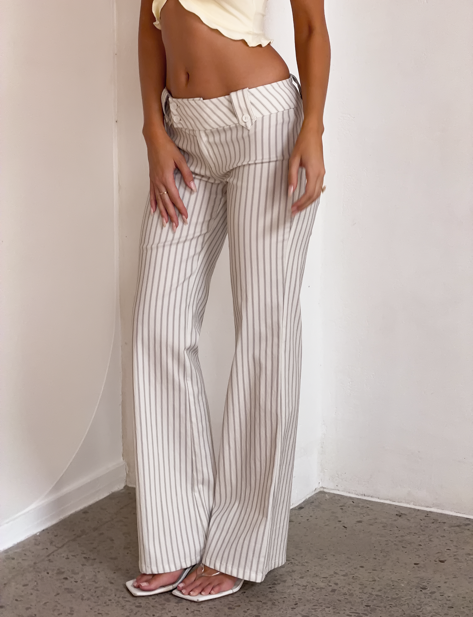A person wears the TIGERMIST KITTIE PANT - IVORY STRIPE, made from stretch suiting fabric with vertical gray stripes, styled with a cropped pale yellow top and white sandals against a plain white wall.