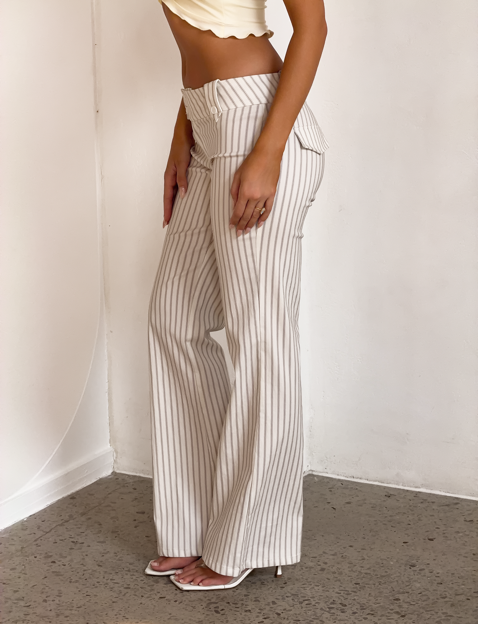 A woman stands sideways wearing TIGERMIST’s KITTIE PANT in ivory stripe, a yellow cropped top with a ruffled hem, and white heeled sandals, posed against a light-colored wall and floor.