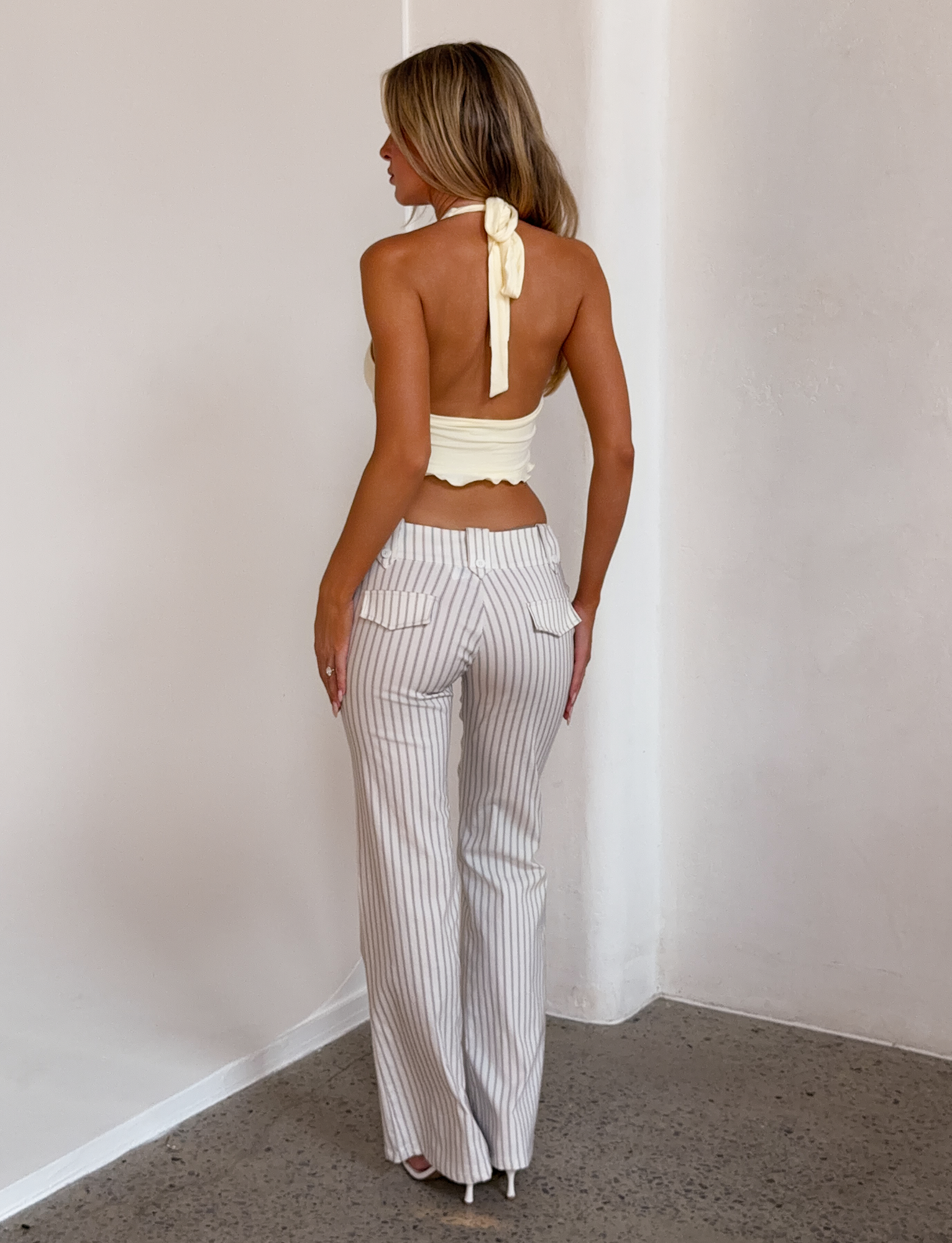 A woman stands indoors facing away, wearing a backless yellow halter top with a bow and TIGERMIST KITTIE PANT - IVORY STRIPE high-waisted flare pants, on a polished concrete floor near a white wall.