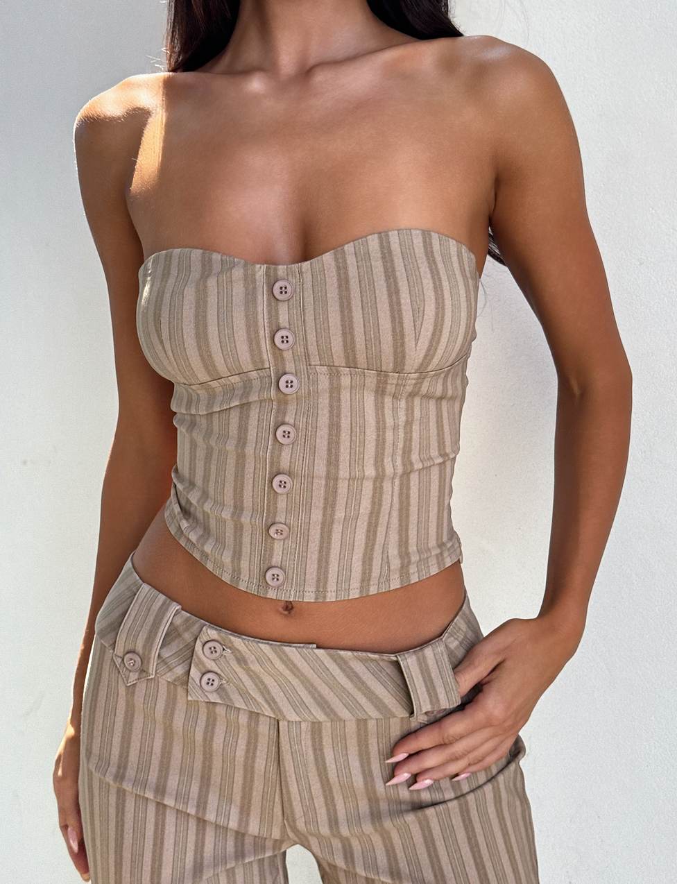 A woman models the TIGERMIST KITTIE STRAPLESS CORSET - TAUPE STRIPE with front buttons, paired with matching high-waisted stretch suiting pants, standing against a plain white background with one hand on her hip.