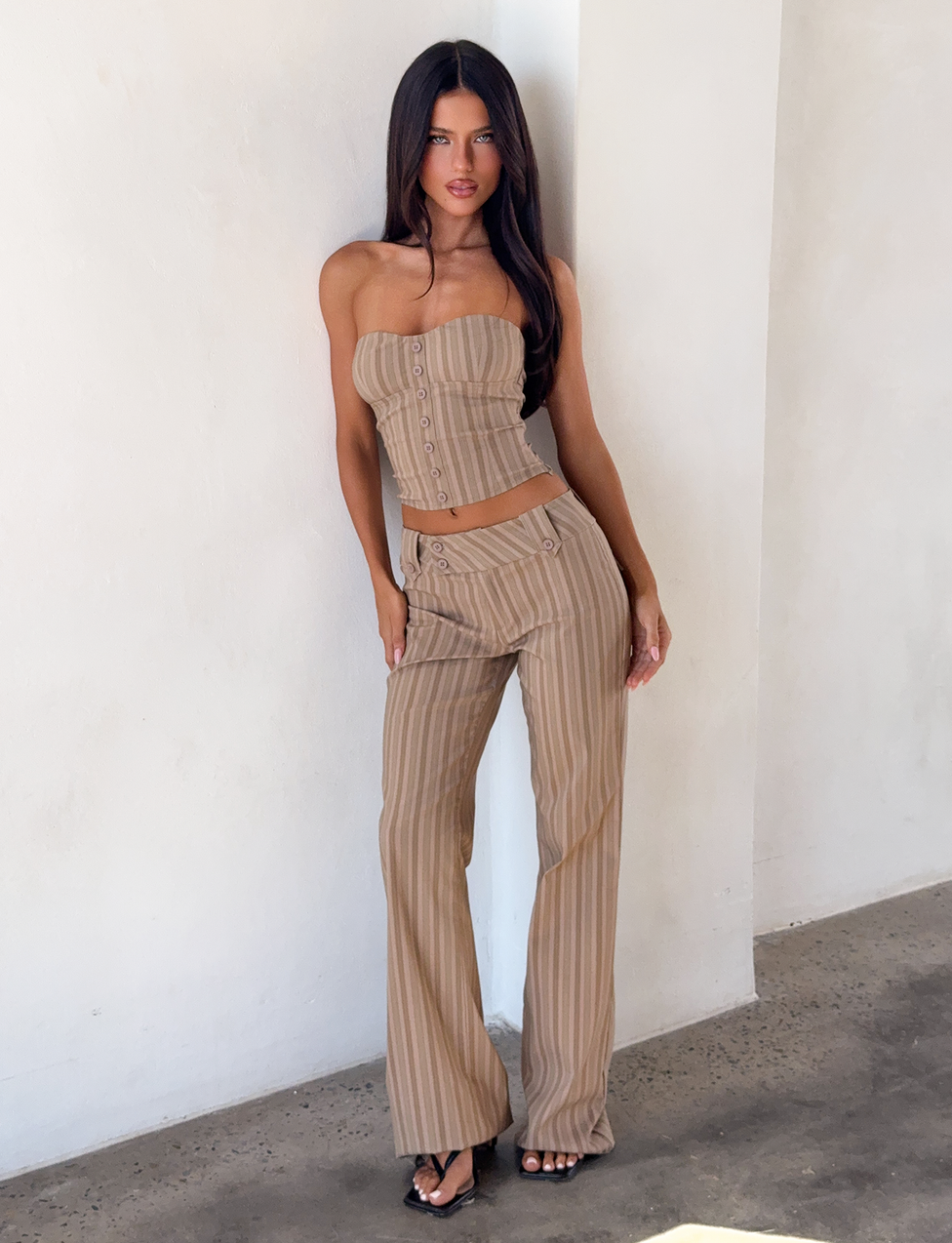 A woman with long dark hair stands against a white wall wearing the TIGERMIST KITTIE PANT - TAUPE STRIPE, a pinstriped stretch suiting pant, paired with a matching strapless top and black heeled sandals.