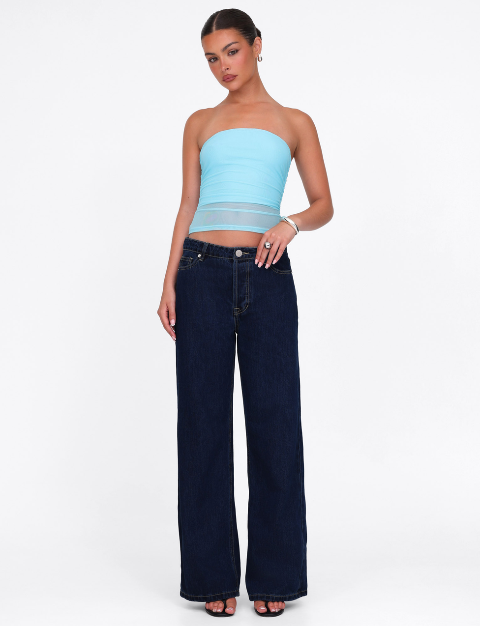 A woman, hair pulled back, poses against a white background wearing the TIGERMIST HARRIETT TUBE TOP in Bright Blue, styled with dark wide-leg jeans, black heels, and silver hoop earrings. One hand rests at her waist.