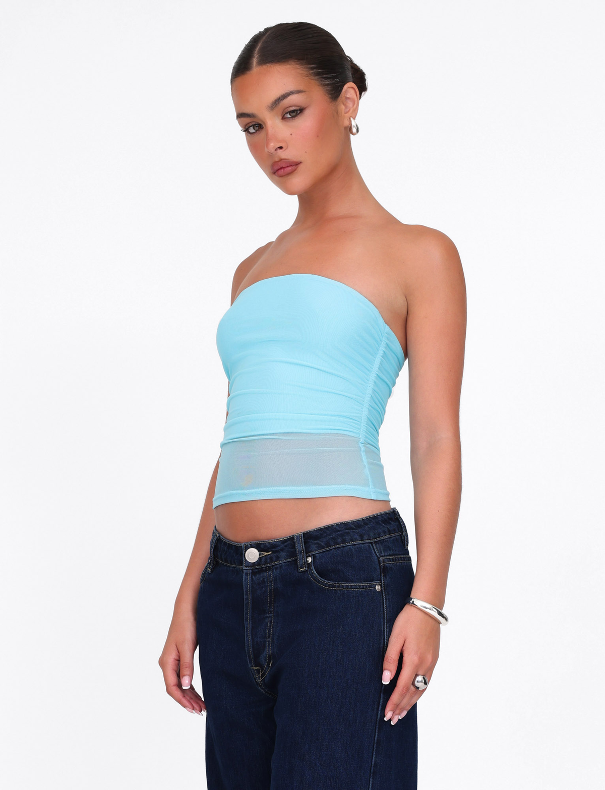 A woman with slicked-back hair wears the TIGERMIST HARRIETT TUBE TOP in ICE BLUE / BRIGHT BLUE and dark blue jeans, accessorized with hoop earrings, a bracelet, and rings, standing against a white background. The top is made from soft mesh fabric.
