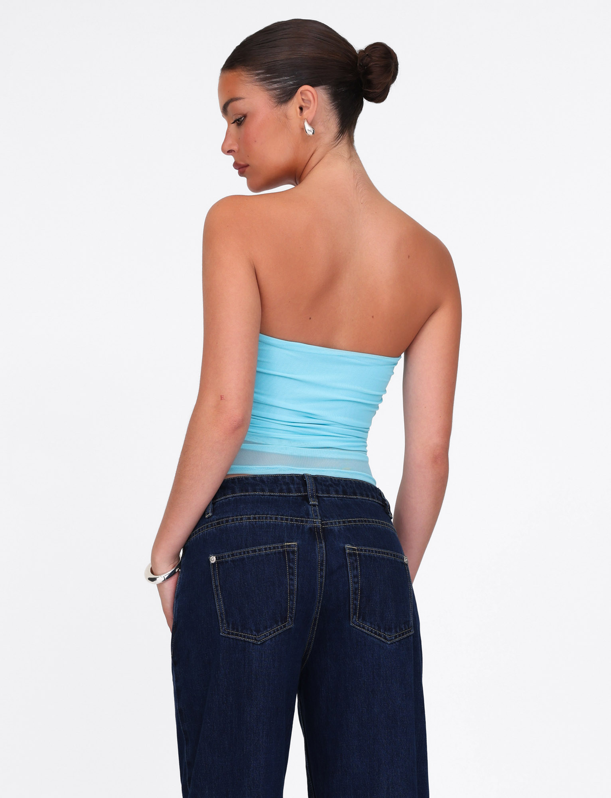 A woman with dark hair in a bun wears the TIGERMIST HARRIETT TUBE TOP in bright blue mesh and dark jeans, standing against a plain white background, looking over her shoulder.