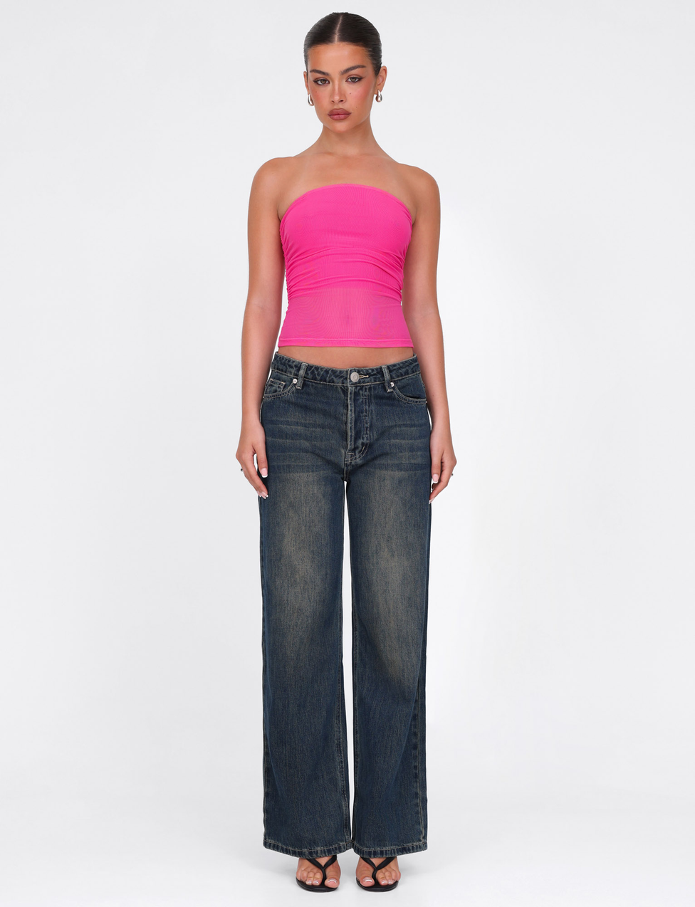 A woman stands against a white background, wearing the TIGERMIST HARRIETT TUBE TOP in pink with loose dark blue jeans and black heeled sandals. Her hair is tied back as she looks straight at the camera.