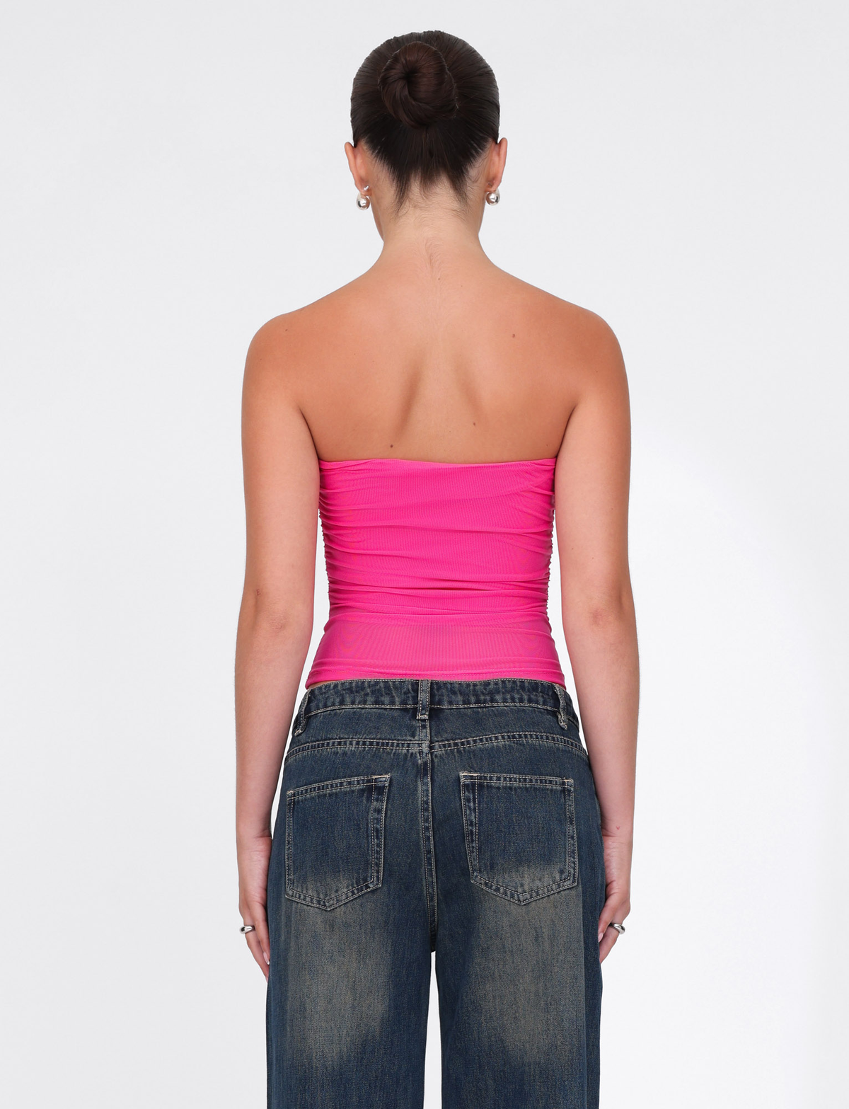 A woman with dark hair in a bun faces away, wearing the TIGERMIST HARRIETT TUBE TOP - PINK and loose blue jeans against a plain white background.