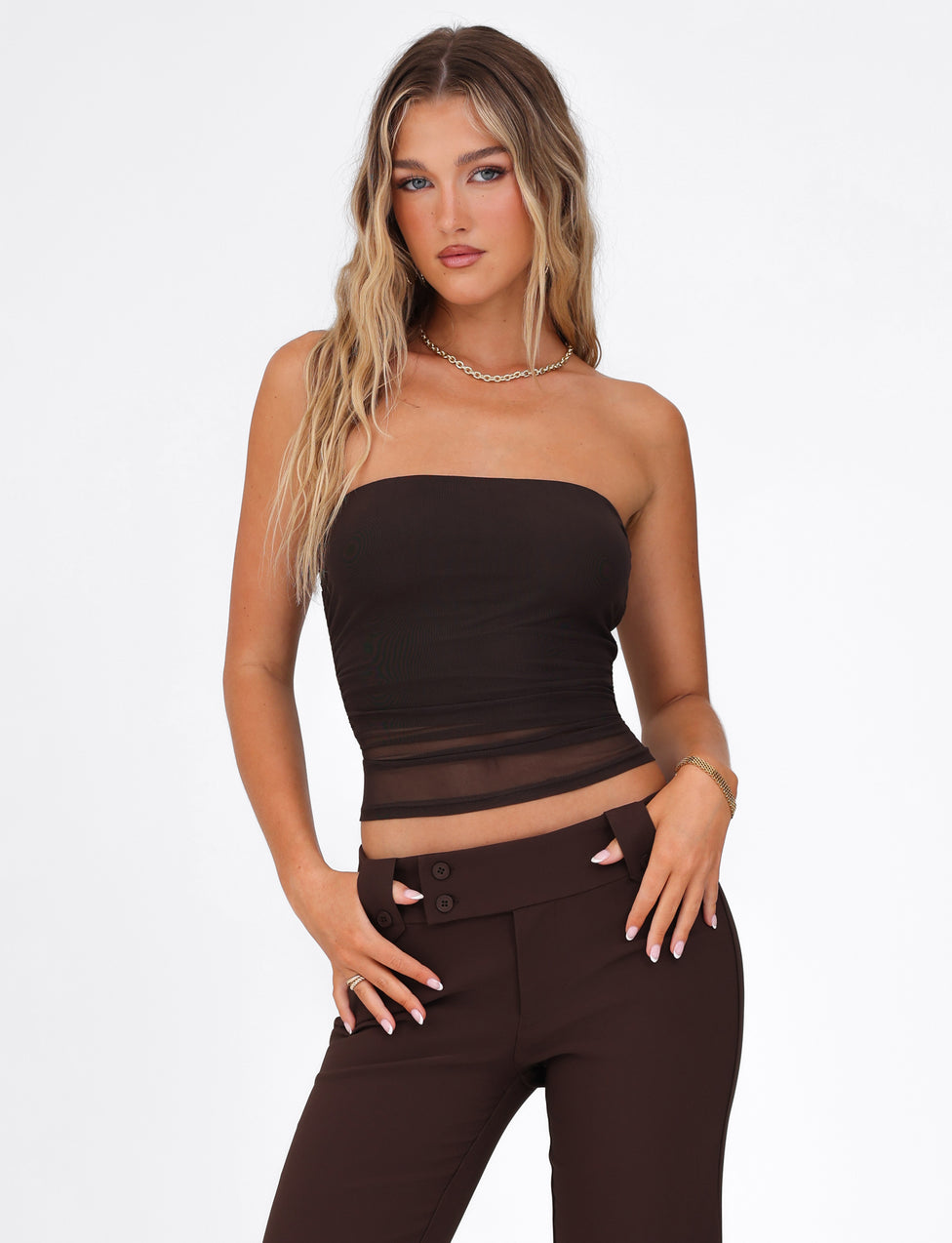 A woman with long wavy hair wears the TIGERMIST HARRIETT TUBE TOP - BROWN : CHOCOLATE, a strapless mesh top, paired with high-waisted dark brown pants. She stands against a white background with one hand in her pocket and a neutral expression.