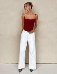 A woman stands against a neutral background wearing a fitted, sleeveless red top with spaghetti straps and the FELIX JEAN - WHITE wide-leg pants from TIGERMIST. Her hair is styled in a casual updo, and she accessorizes with a necklace, bracelet, and pointed shoes.