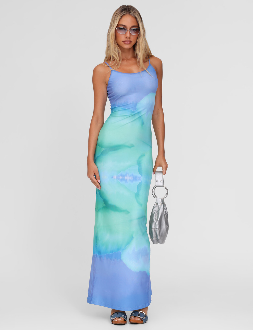 A woman in the TIGERMIST ARDON MAXI DRESS - BLUE : MACRO FLORAL : BLUE HIBISCUS, blue heeled sandals, sunglasses, and a silver handbag poses against a plain white background.
