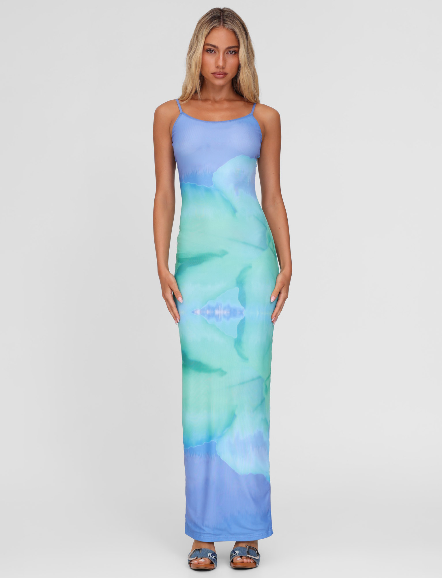 A woman with long blonde hair faces forward in the TIGERMIST ARDON MAXI DRESS - BLUE HIBISCUS, a blue mesh dress with macro floral print. She pairs it with blue open-toe sandals, standing against a plain white background.