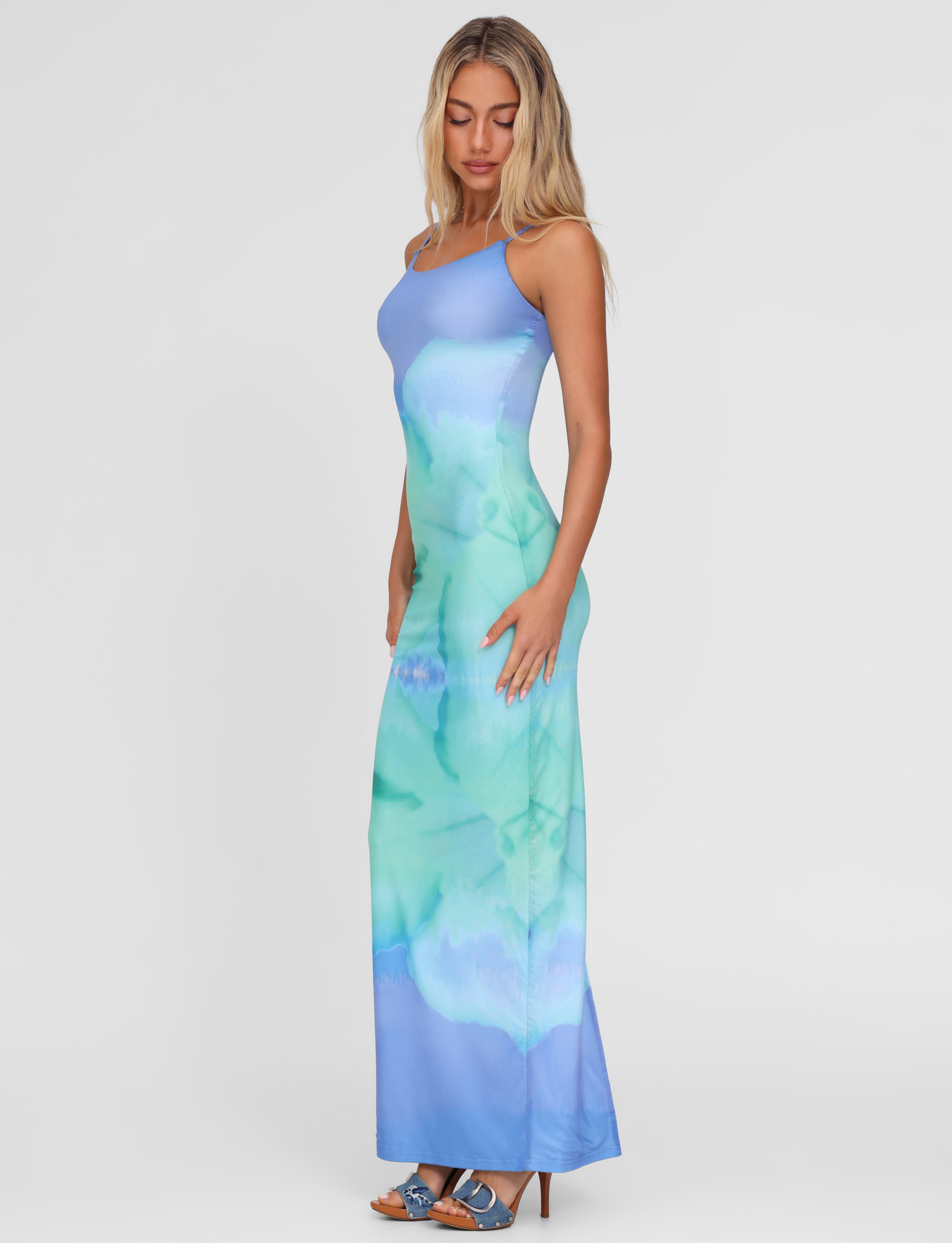 A woman stands sideways in heels against a white background, wearing the TIGERMIST ARDON MAXI DRESS - BLUE, featuring a sleeveless, form-fitting silhouette and blue macro floral hibiscus print in high-stretch fabric.