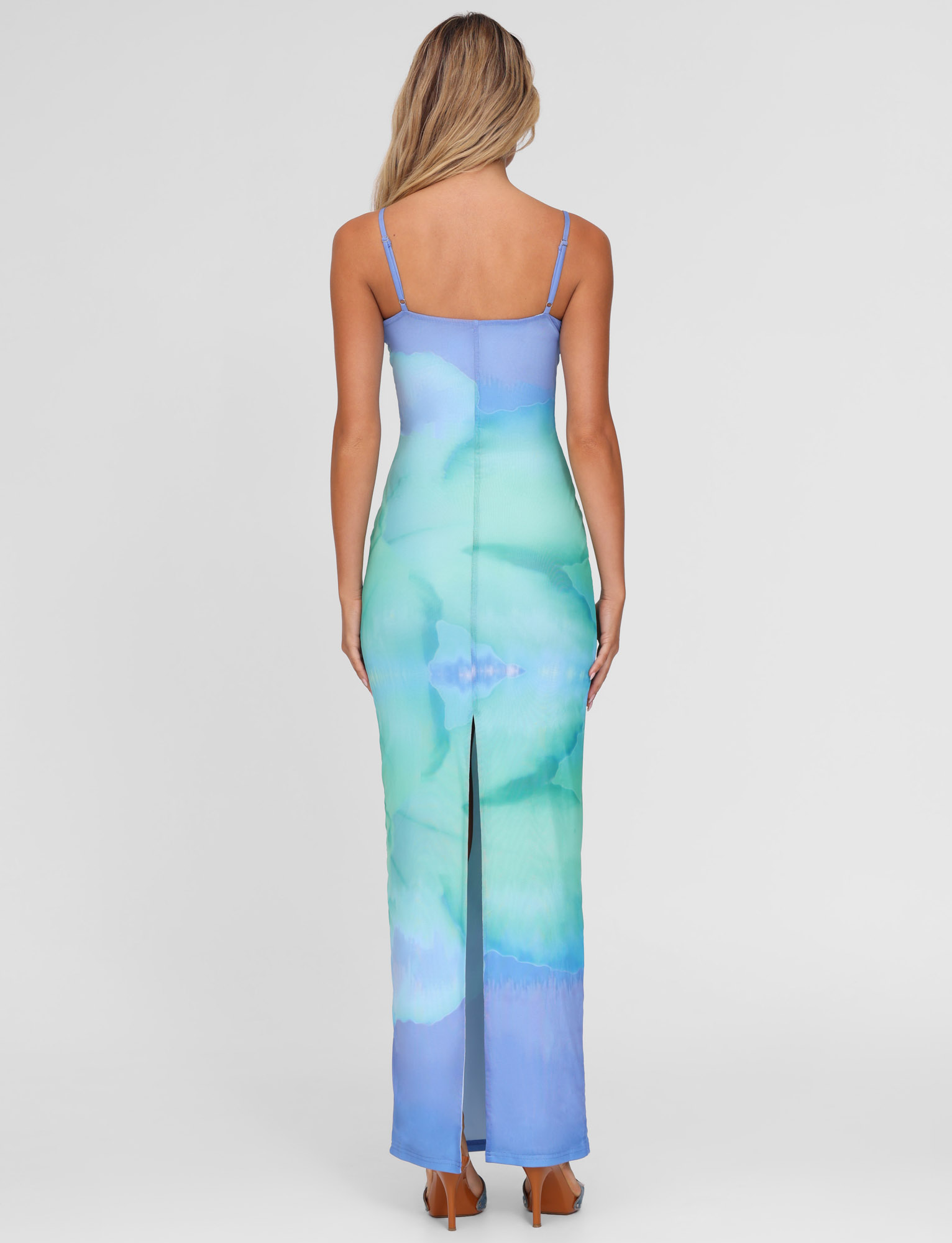 A woman with long, wavy blonde hair is seen from behind wearing the TIGERMIST ARDON MAXI DRESS in Macro Floral Blue Hibiscus print, featuring thin straps and a back slit, standing against a plain white background.