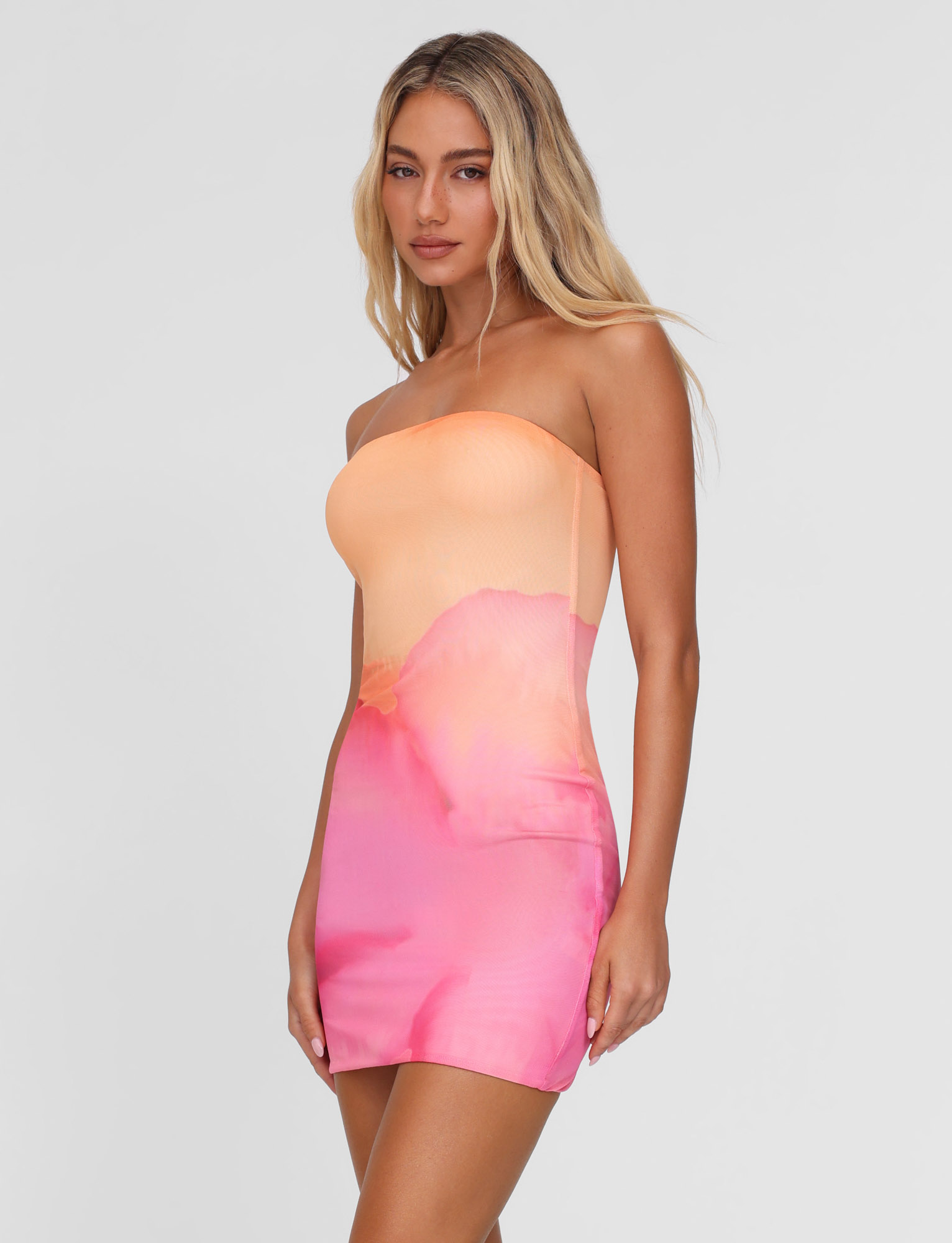 A woman with long blonde hair wears the TIGERMIST CHER MINI DRESS in Macro Floral Pink Hibiscus, featuring a strapless, bodycon fit, standing against a plain light gray background.
