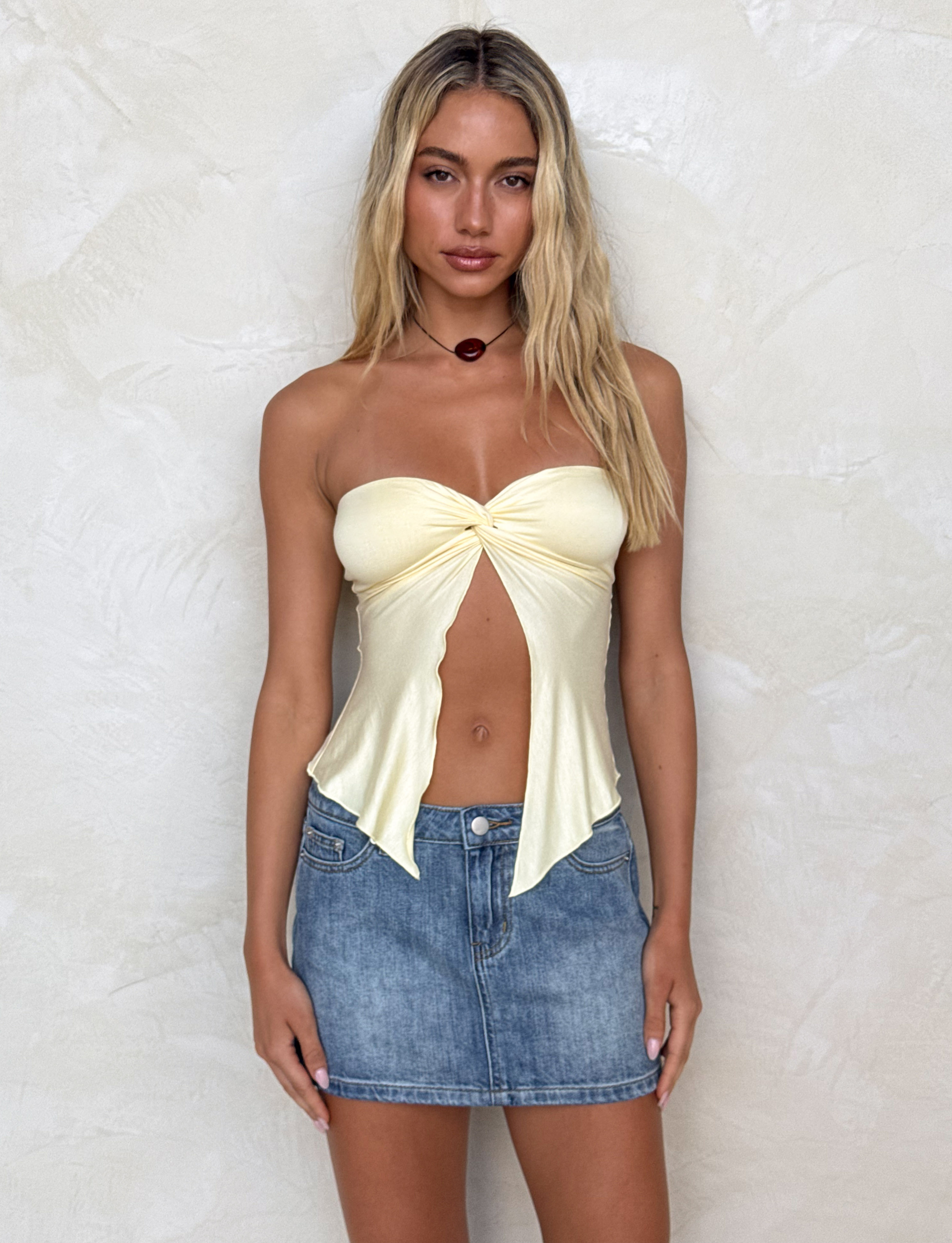 A woman with long blonde hair stands against a light textured wall, wearing the TIGERMIST CHARMAINE TOP in lemon and a blue denim mini skirt. She looks directly at the camera with a neutral expression.