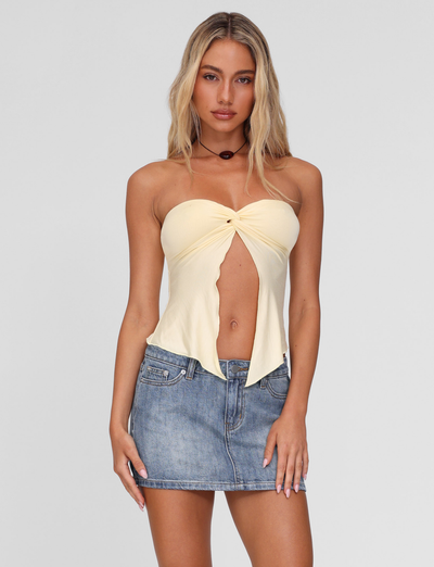 A woman with long blonde hair wears the TIGERMIST CHARMAINE TOP in lemon and a short denim skirt, standing against a plain light gray background with a neutral expression.