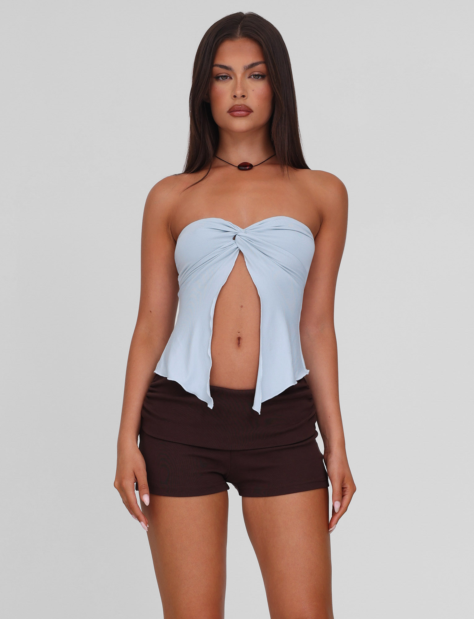 A woman with long dark hair wears the TIGERMIST CHARMAINE TOP in baby blue, a strapless style with twist detail, paired with black shorts. She stands facing forward against a plain light gray background.