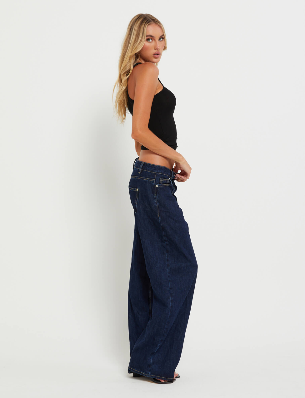 A woman with long blonde hair stands sideways against a white background, wearing a black tank top and TIGERMIST FELIX JEAN - DARK DENIM wide-leg jeans in an oversized fit.