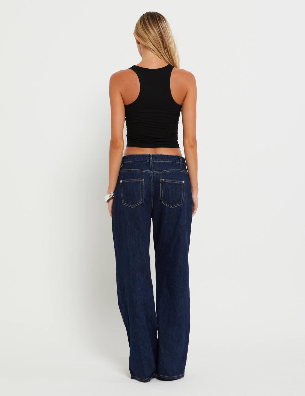 A woman with long blonde hair faces away, wearing a black sleeveless crop top and TIGERMIST FELIX JEAN - DARK DENIM wide leg pants, made from stretch denim, against a plain white background.