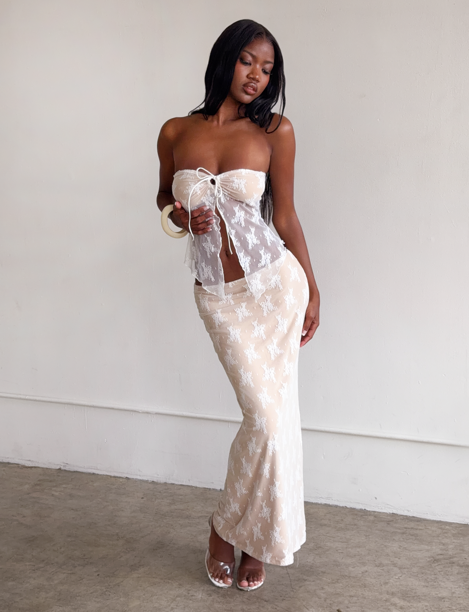 A woman poses indoors against a light wall wearing the Tiger Mist USA CL - KAROLINA TOP, a sheer white lace strapless top paired with a matching long skirt, styled with white high-heeled sandals and large hoop earrings.