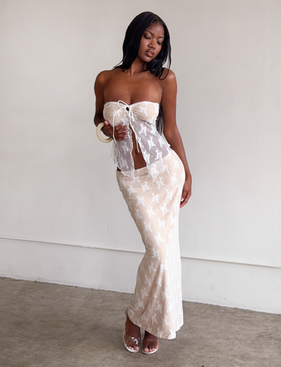 A woman with long dark hair stands indoors, wearing a sheer white strapless top and the KAROLINA SKIRT - WHITE LACE by TIGERMIST—a pull-on skirt with a delicate floral pattern. She holds a large bangle and gazes down, one knee slightly bent.