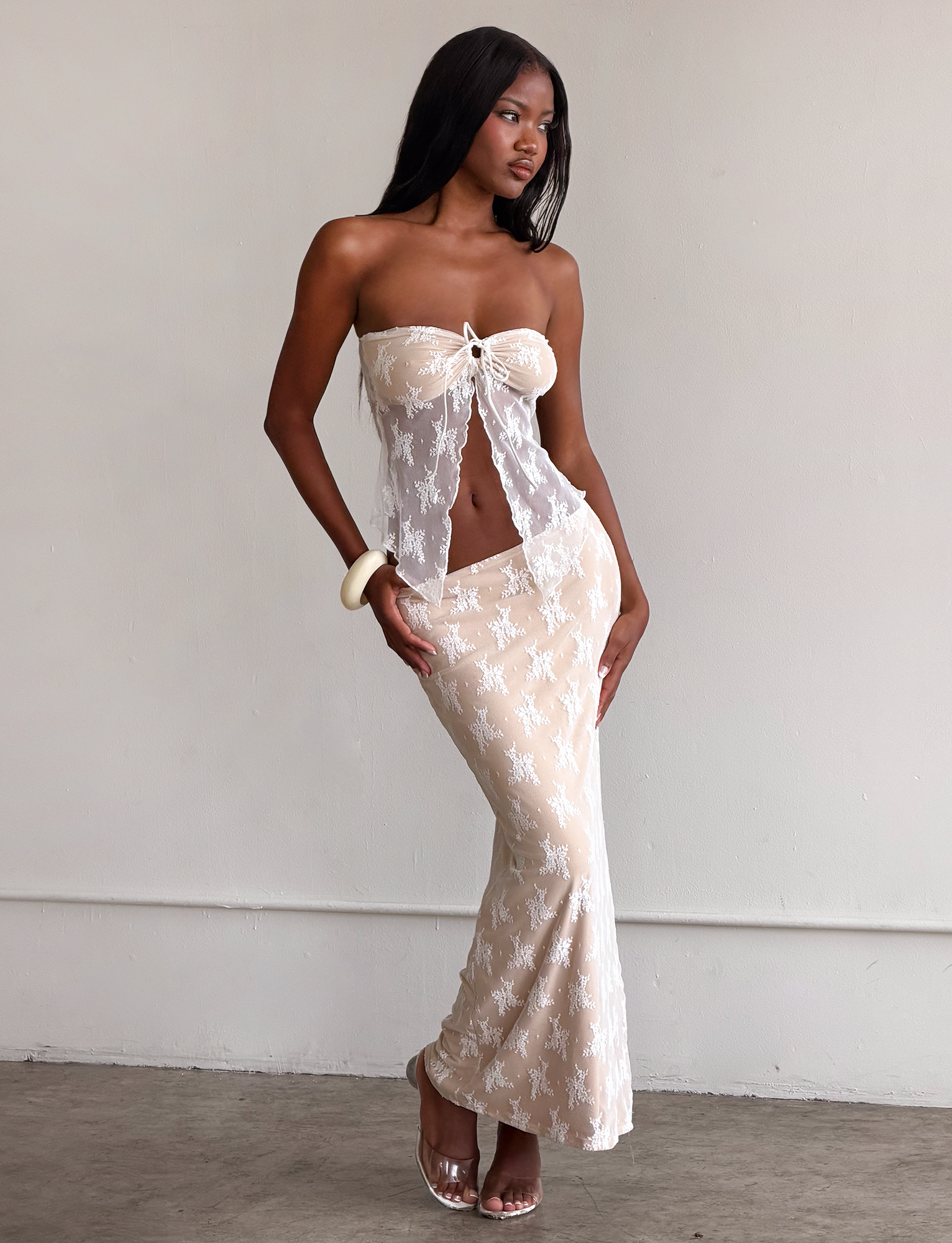 A woman poses indoors wearing TIGERMIST’s KAROLINA SKIRT - WHITE in white lace, styled over a nude underlayer and paired with a sheer, strapless top. She accessorizes with clear heels and a white bracelet.