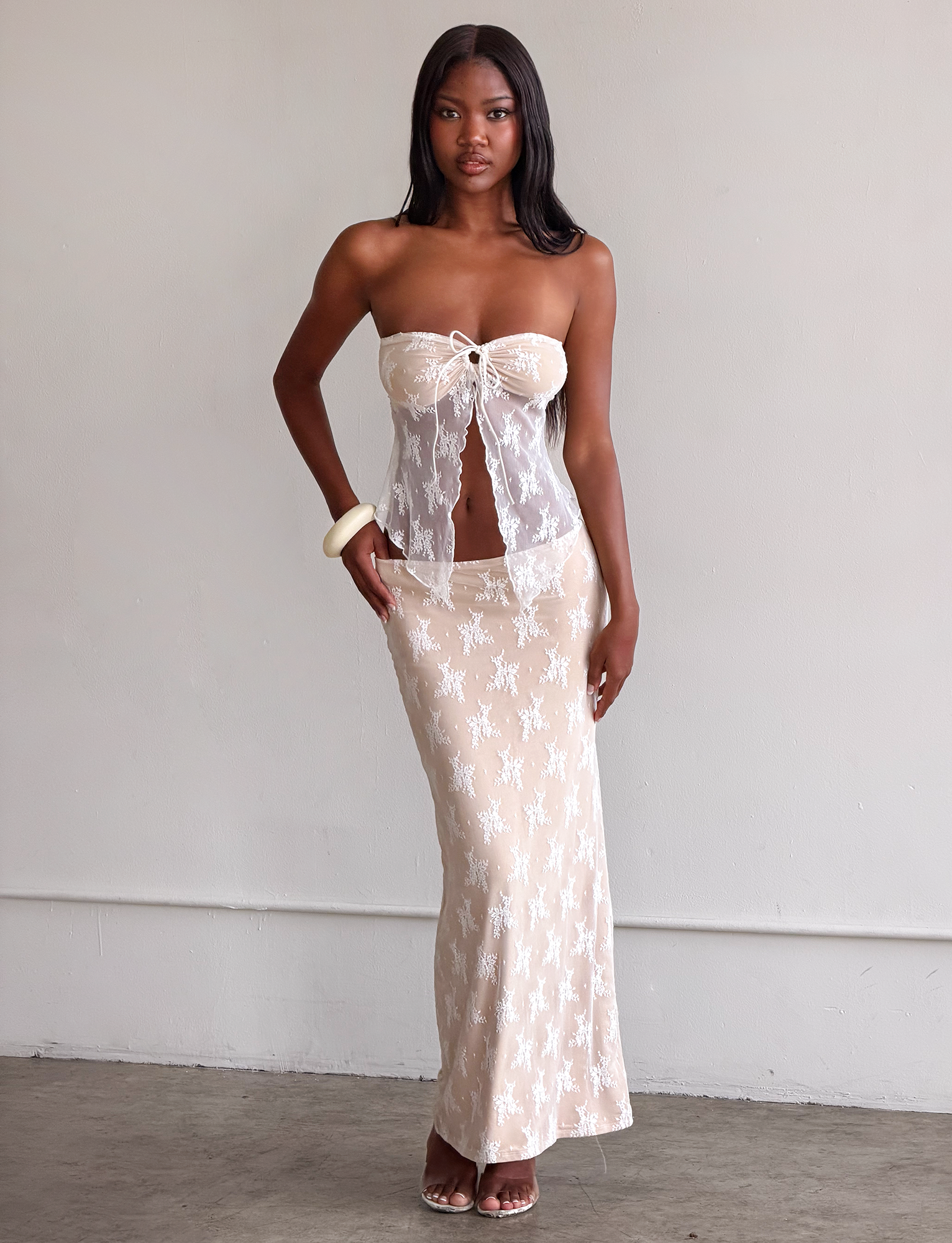 A woman with long straight hair and a neutral expression wears the TIGERMIST KAROLINA TOP – WHITE, a sheer white lace strapless top with a subtle bust cutout, paired with a matching lace skirt, white bracelet, and sandals against a light background.